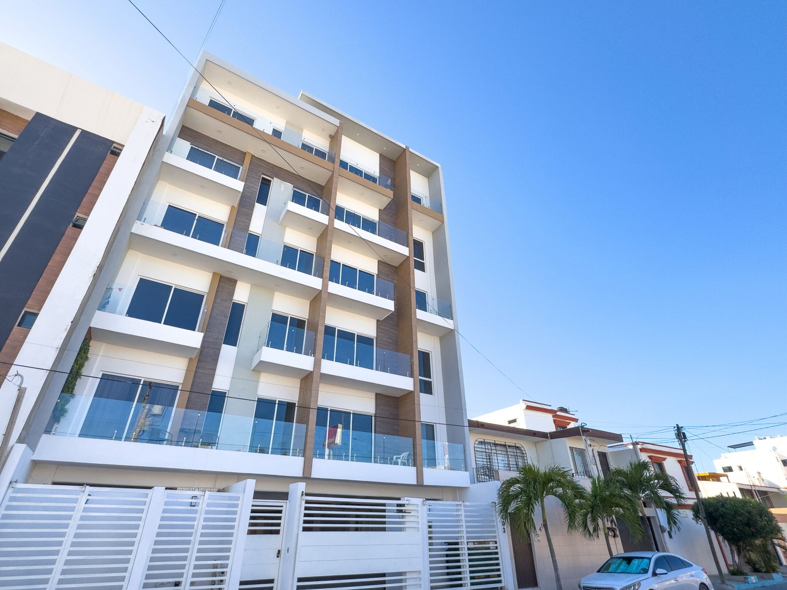 Edificio de departamentos de varios niveles con portón de seguridad blanco y palmeras decorativas en la entrada.