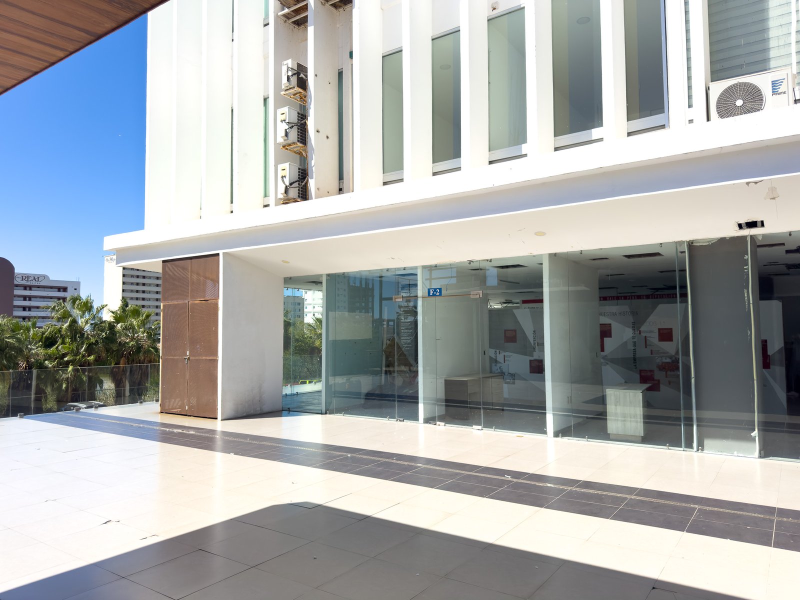 Vista frontal desde el pasillo exterior del local F2 en Laguna Plaza, destacando ventanales de piso a techo y fachada blanca.