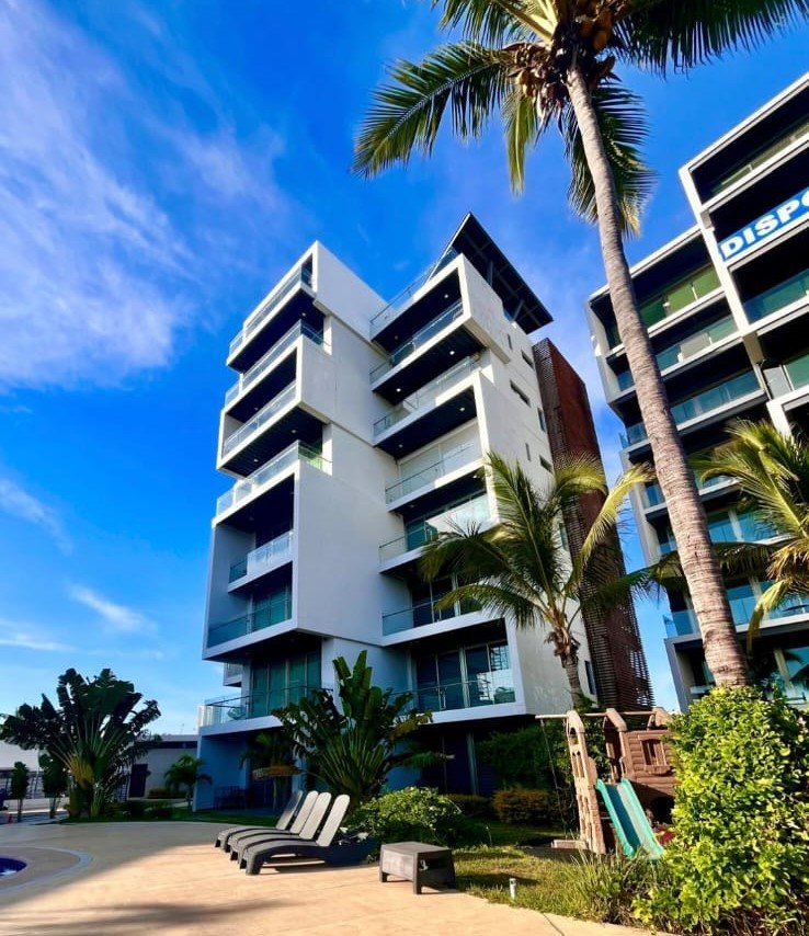 Fachada moderna de los departamentos Manzara Loft con balcones amplios, palmeras y cielo despejado.